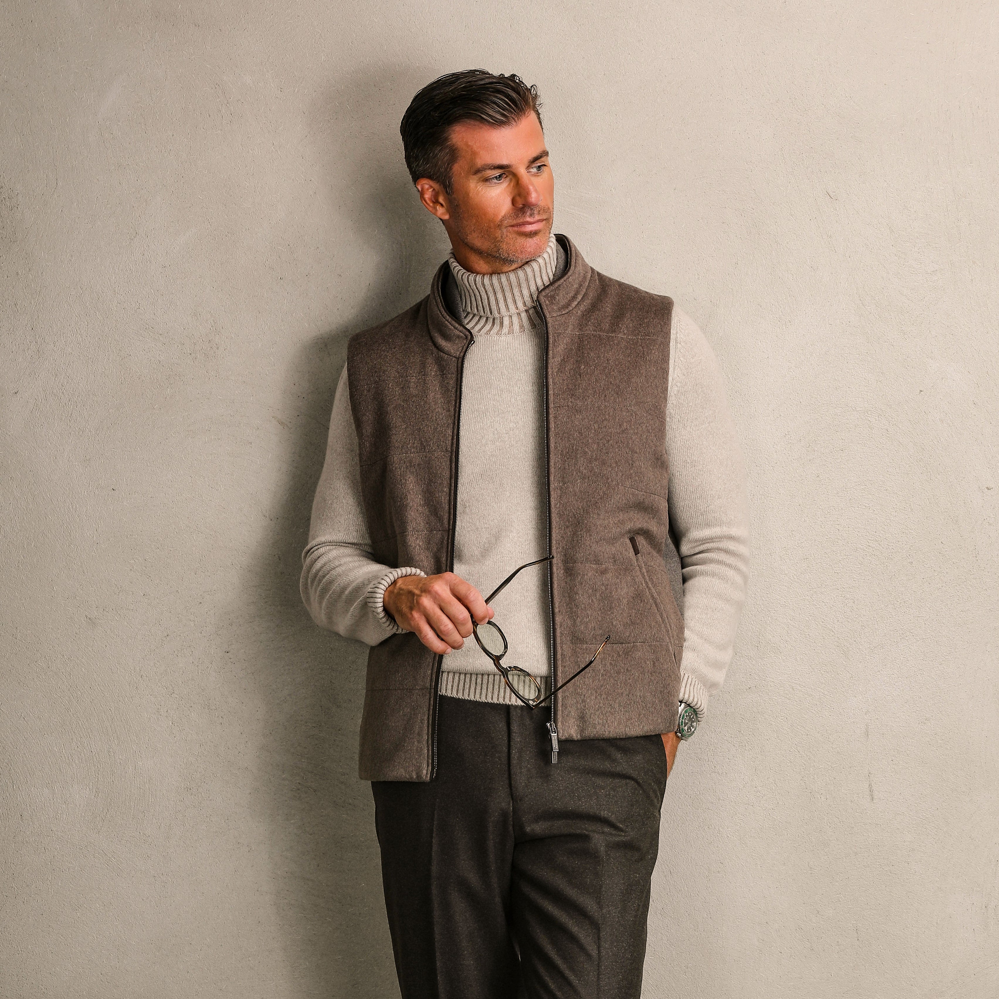 A man leans against a plain wall, wearing a light turtleneck, a Dark Taupe Water Repellent Cashmere Gilet by Maurizio Baldassari, and holding eyeglasses in one hand.