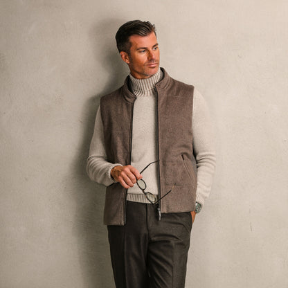 A man leans against a plain wall, wearing a light turtleneck, a Dark Taupe Water Repellent Cashmere Gilet by Maurizio Baldassari, and holding eyeglasses in one hand.