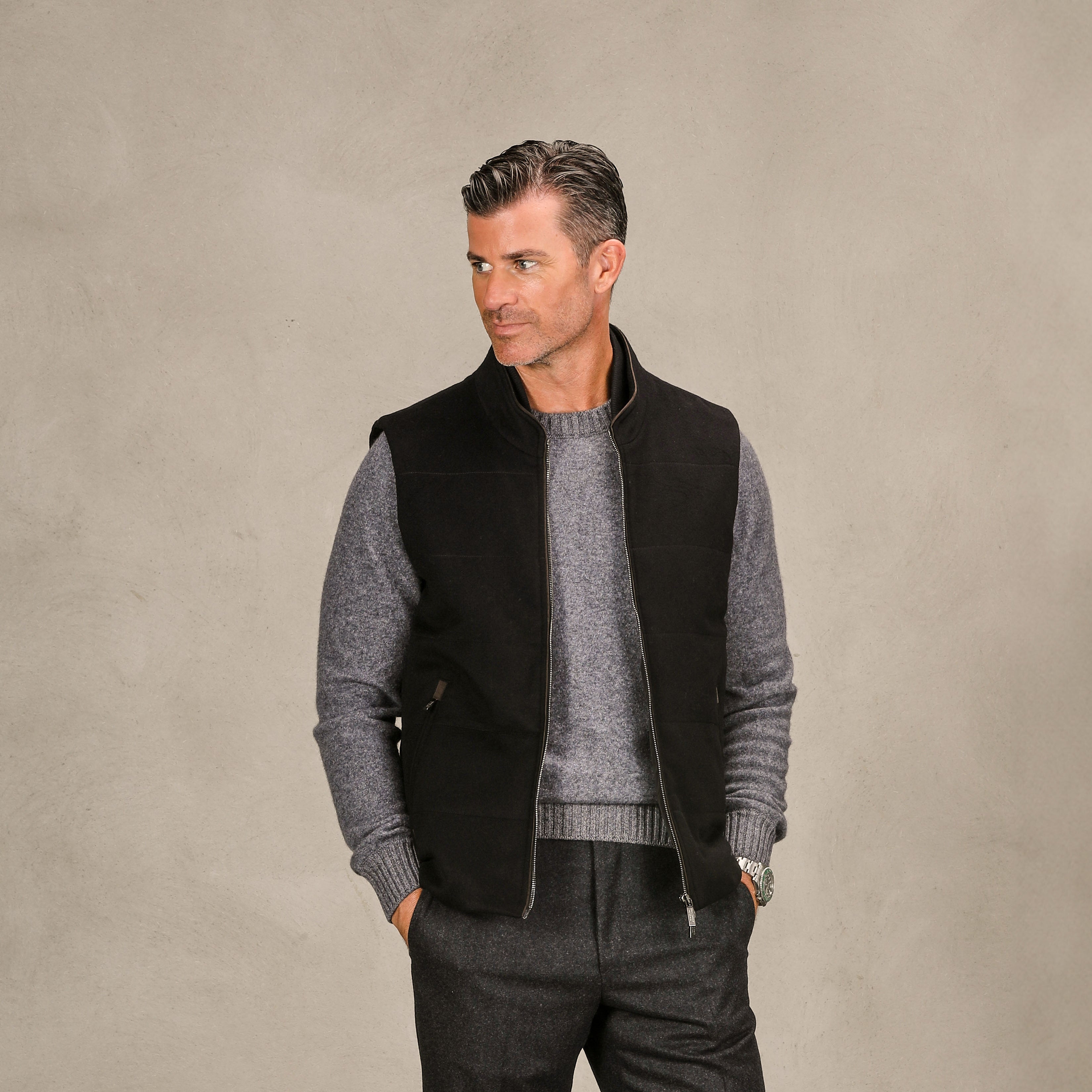 A man in a dark vest over a Gran Sasso Grey Felted Cashmere Crew Neck Sweater and trousers stands against a plain background, looking to the side.