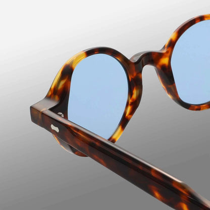 Close-up of TBD Eyewear’s Giotto Eco Spotted Havana | Blue round eyeglass frames with light blue lenses, highlighting the glossy tortoiseshell pattern on the front and side against a smooth gray gradient background. , available at Richard Gelding Mayfair