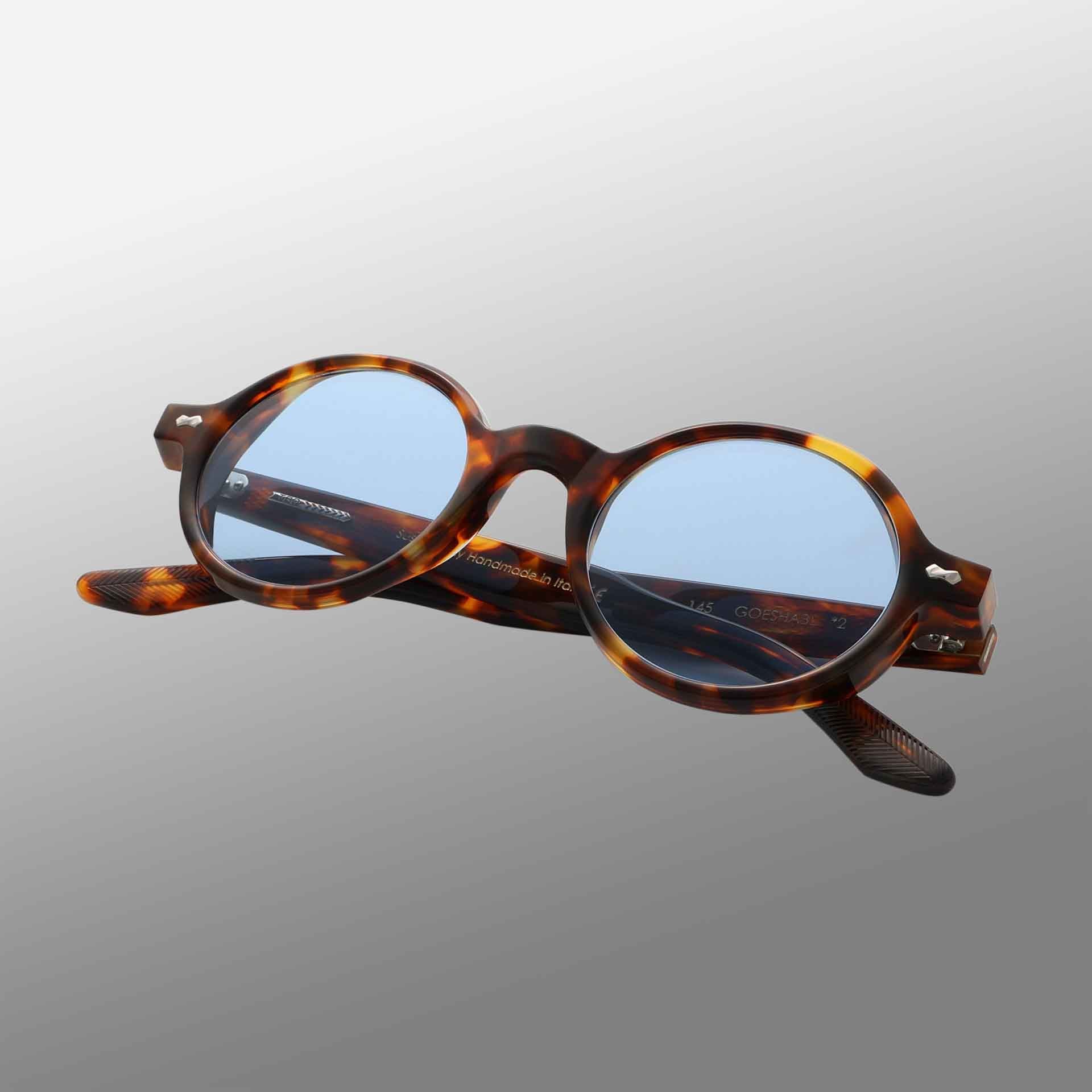 The Giotto Eco Spotted Havana | Blue eyeglasses by TBD Eyewear feature thick tortoiseshell-patterned frames with light blue lenses, metal hinge details, and engraved text inside the slightly open arms, set against a gray gradient background. , available at Richard Gelding Mayfair