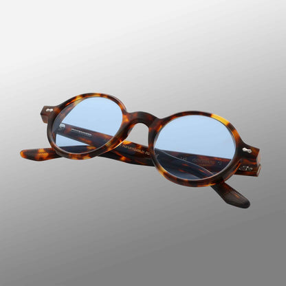 The Giotto Eco Spotted Havana | Blue eyeglasses by TBD Eyewear feature thick tortoiseshell-patterned frames with light blue lenses, metal hinge details, and engraved text inside the slightly open arms, set against a gray gradient background. , available at Richard Gelding Mayfair