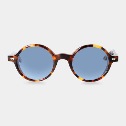 The Giotto Eco Spotted Havana | Blue sunglasses by TBD Eyewear feature round blue lenses and a tortoiseshell-patterned frame, displayed front-facing on a white background with metallic accents on the temples near the hinges. , available at Richard Gelding Mayfair