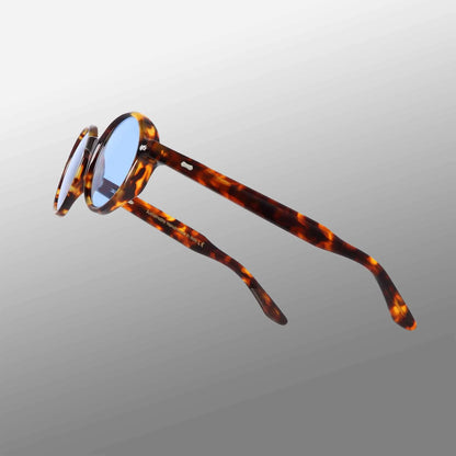 The Giotto Eco Spotted Havana | Blue sunglasses by TBD Eyewear feature blue lenses and tortoiseshell-patterned frames and arms, shown angled on a white-to-gray gradient background with open arms highlighting the spotted amber and brown design. , available at Richard Gelding Mayfair