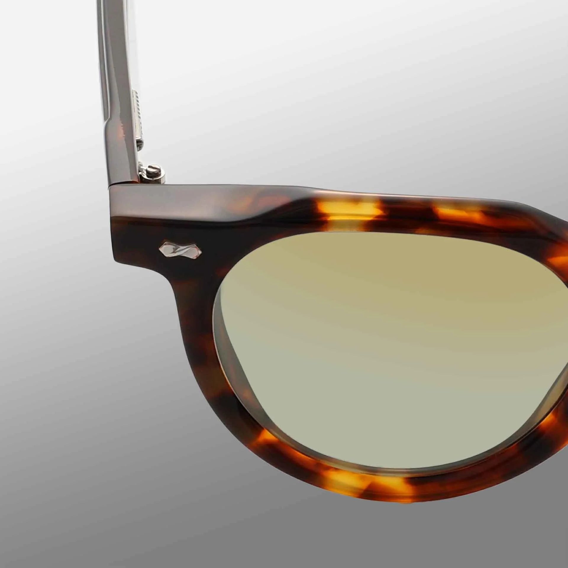 Close-up of the upper left corner of TBD Eyewear's Hem Eco Spotted Havana | Mirror Green sunglasses, showing brown tortoiseshell frame, light yellow-tinted lens, metal hinge, and part of the arm against a gradient gray background. , available at Richard Gelding Mayfair