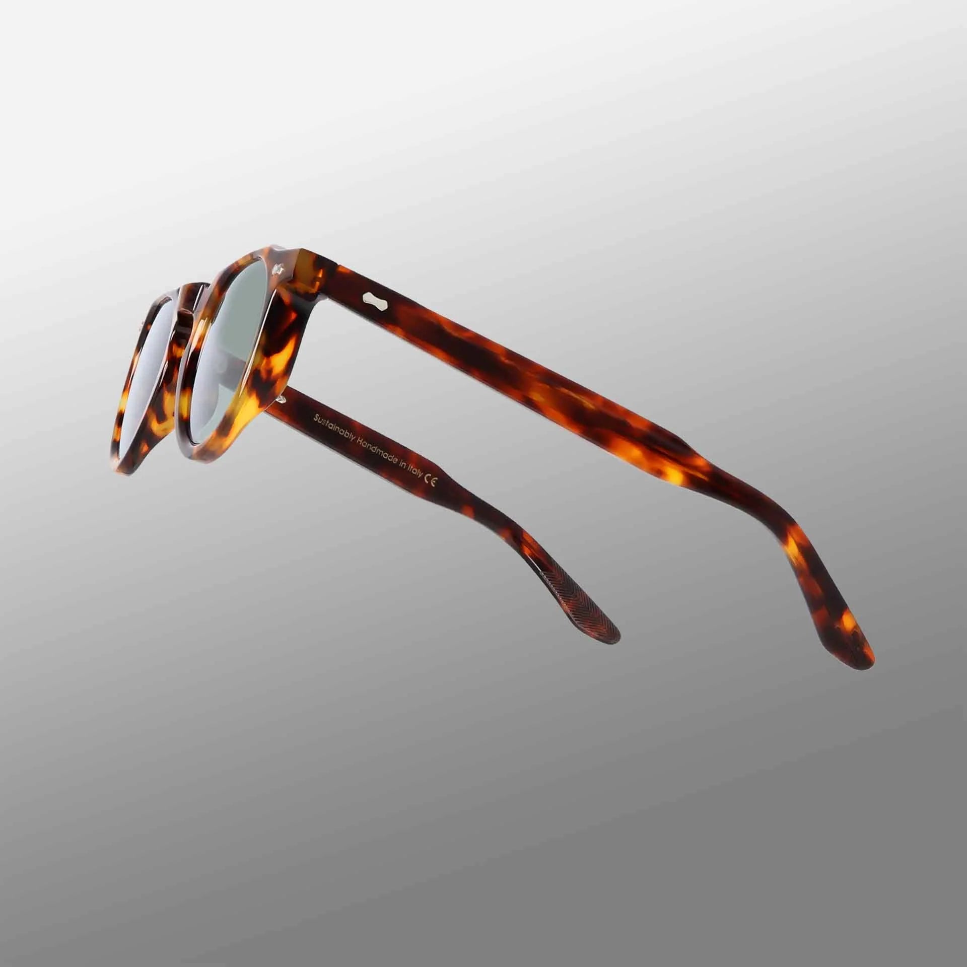 The Hem Eco Spotted Havana | Mirror Green sunglasses by TBD Eyewear are displayed at an angle, showcasing their brown and amber marbled frames with slender arms against a sleek white-to-gray gradient background. , available at Richard Gelding Mayfair