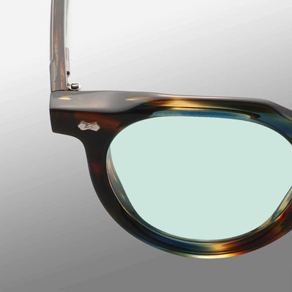 Close-up of the top left of Hem River | Light Green eyeglasses by TBD Eyewear. Features a tortoiseshell frame, light green lens, and a small silver emblem on the temple. Background fades smoothly from light gray to dark gray. , available at Richard Gelding Mayfair
