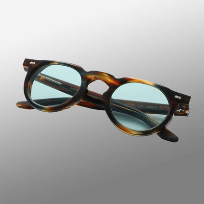 The Hem River | Light Green by TBD Eyewear, featuring thick round tortoiseshell frames and light green-tinted lenses, is displayed against a smooth grey gradient with partially open, overlapping temples and visible hinge details. , available at Richard Gelding Mayfair