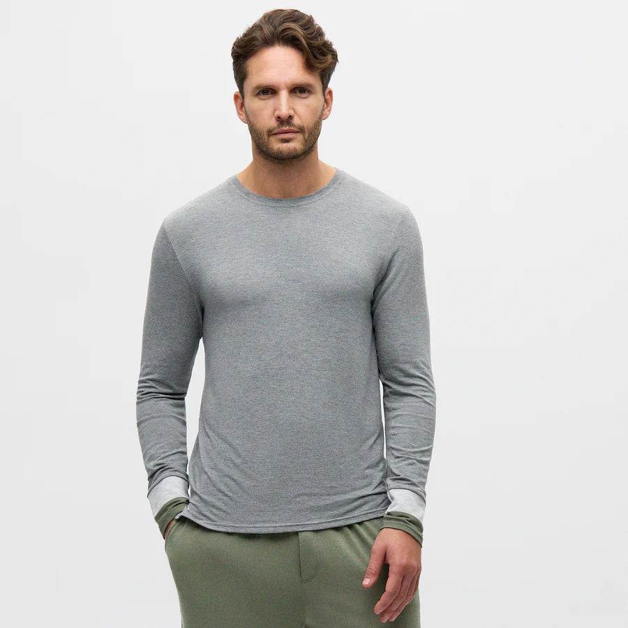 A man with short, wavy brown hair and a trimmed beard stands facing forward, hands in the pockets of olive green pants. He wears a fitted, long-sleeve, light gray shirt. The background is plain white, giving a minimal and clean look. , available at Richard Gelding Mayfair