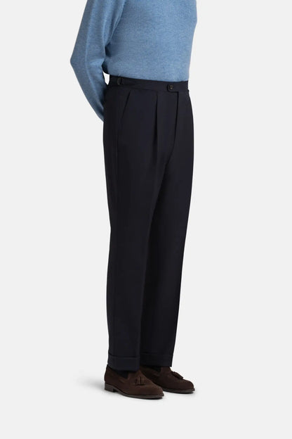 A person wearing Cavour Navy Blue Wide Leg Pleated Twist Wool Trousers with a high-waisted cut and side tab adjusters, paired with a light blue long-sleeve sweater and dark brown suede tassel loafers against a plain, light-colored background. , available at Richard Gelding Mayfair