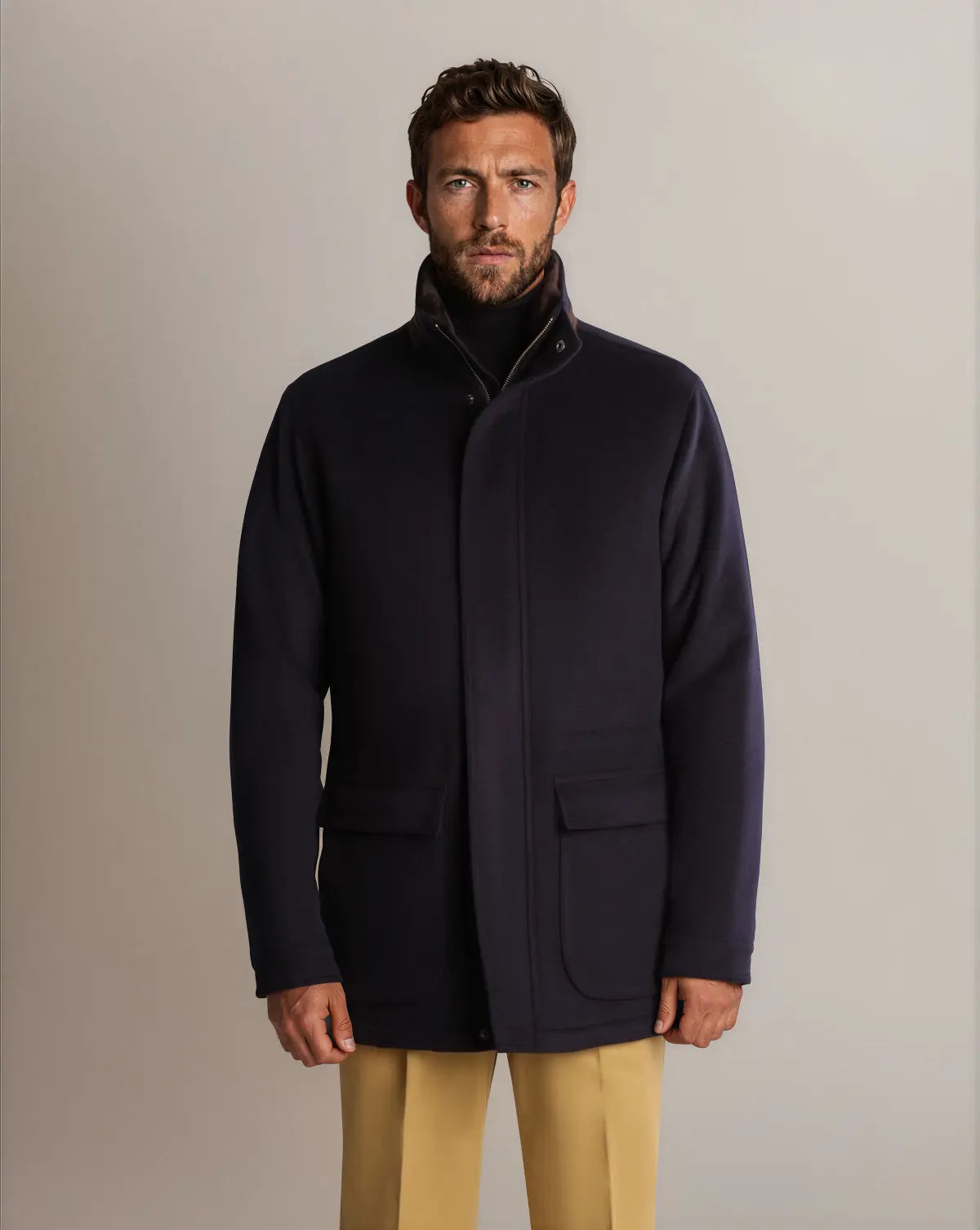 A man with short brown hair and a beard wears a navy Cavour Winter Car Coat with Removable Cashmere Collar over a dark turtleneck and tan trousers against a plain, light-grey background. , available at Richard Gelding Mayfair