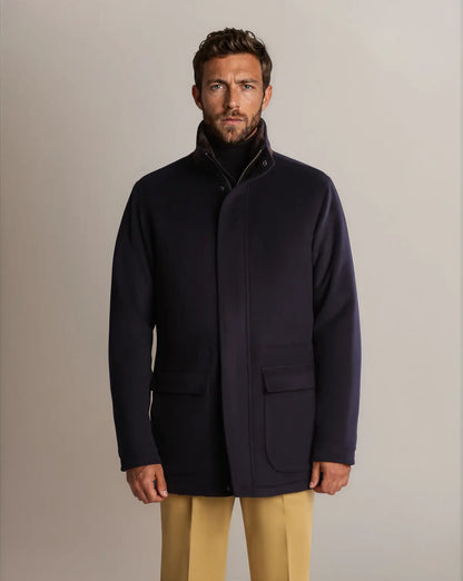 A man with short brown hair and a beard wears a navy Cavour Winter Car Coat with Removable Cashmere Collar over a dark turtleneck and tan trousers against a plain, light-grey background. , available at Richard Gelding Mayfair
