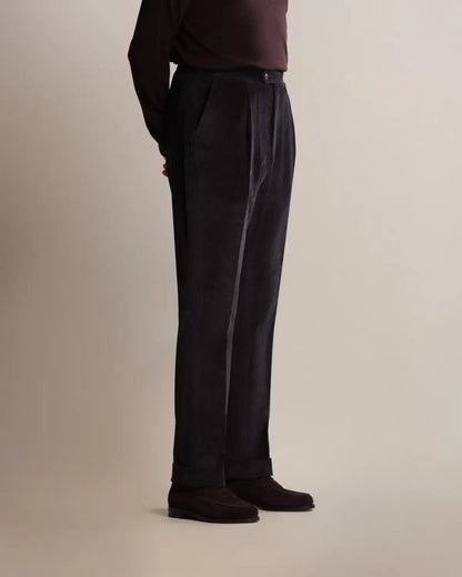 A person viewed from the side wears Cavour Navy Wide Leg Pleated Corduroy Trousers with a wide-leg, pleated fit and cuffed hems, paired with a dark brown long-sleeved shirt and matching dark suede loafers against a plain light background. , available at Richard Gelding Mayfair