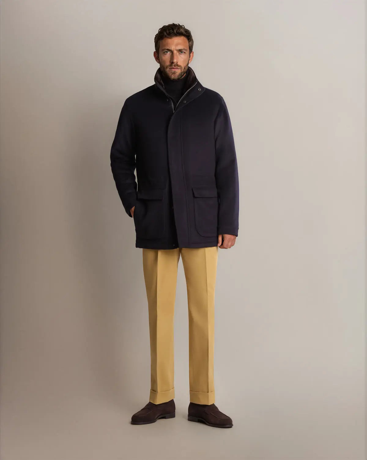 A man wears a Cavour Navy Winter Car Coat with Removable Cashmere Collar over a black turtleneck, mustard-yellow trousers, and dark brown suede loafers against a solid, neutral gray background. , available at Richard Gelding Mayfair