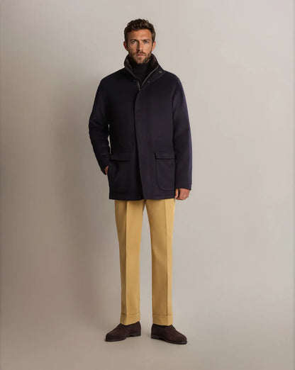A man wears a Cavour Navy Winter Car Coat with Removable Cashmere Collar over a black turtleneck, mustard-yellow trousers, and dark brown suede loafers against a solid, neutral gray background. , available at Richard Gelding Mayfair