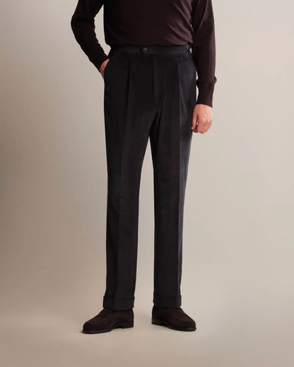 A man stands wearing navy blue Cavour Navy Wide Leg Pleated Corduroy Trousers with double pleats and a wide-leg cut, paired with a long-sleeved dark maroon sweater and brown suede loafers against a plain light gray background. , available at Richard Gelding Mayfair