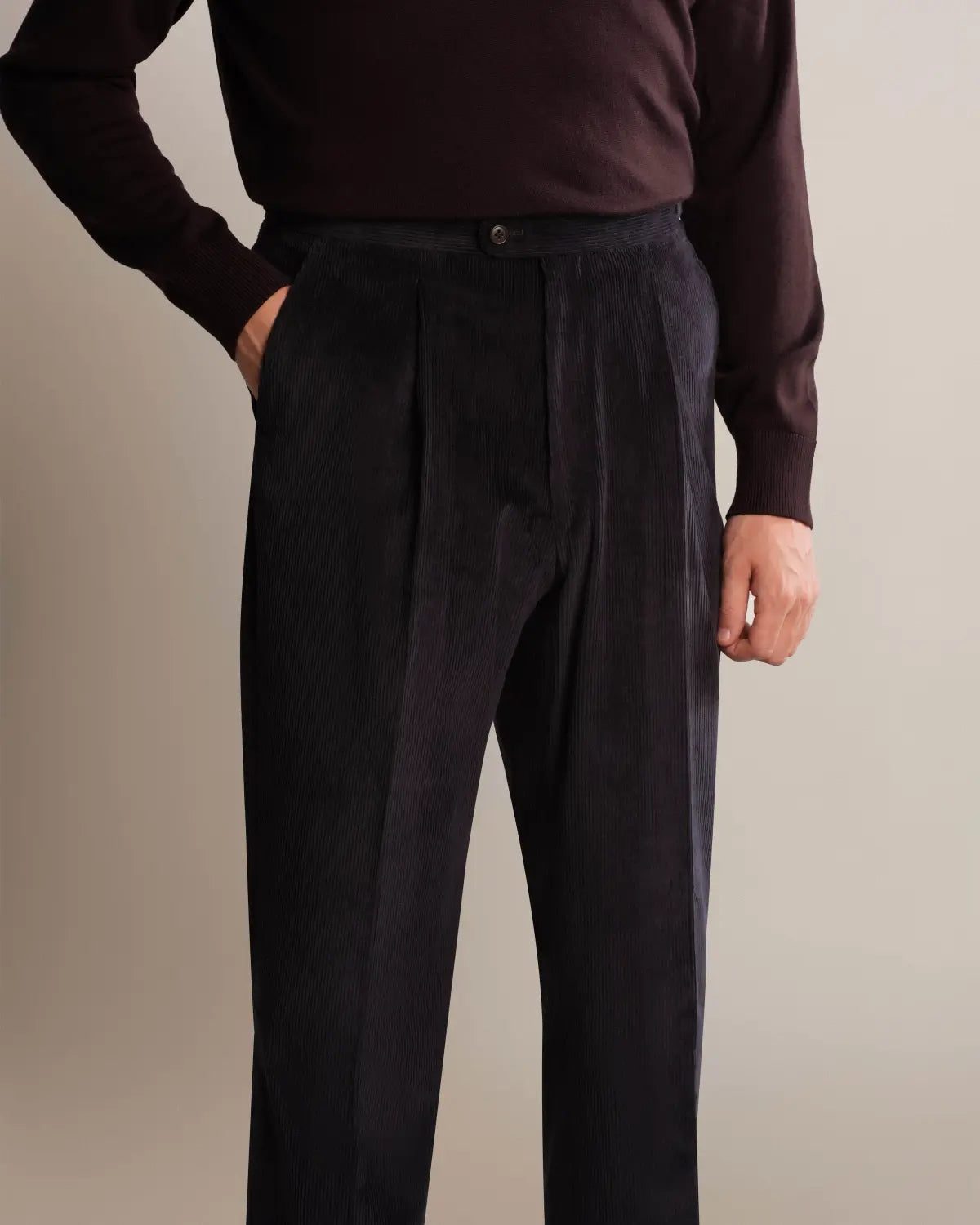 A person wears Cavour Navy Wide Leg Pleated Corduroy Trousers with a dark brown long-sleeved sweater against a neutral background; the trousers feature a classic button closure and a vertical ribbed texture. , available at Richard Gelding Mayfair