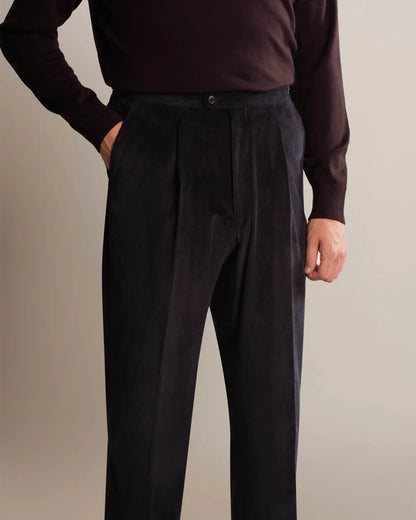 A person wears Cavour Navy Wide Leg Pleated Corduroy Trousers with a dark brown long-sleeved sweater against a neutral background; the trousers feature a classic button closure and a vertical ribbed texture. , available at Richard Gelding Mayfair