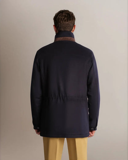 A man seen from the back wears a Navy Winter Car Coat with Removable Cashmere Collar over tan trousers; the mid-length jacket features a cinch at the waist, set against a plain, neutral light gray background. , available at Richard Gelding Mayfair