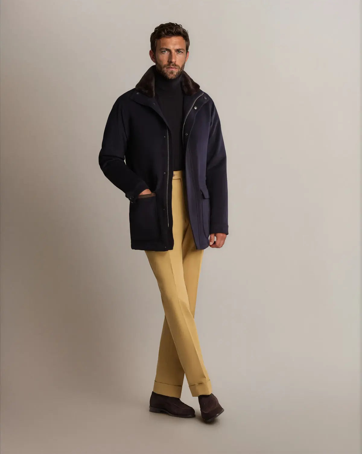 A man wearing a Cavour Navy Winter Car Coat with Removable Cashmere Collar over a black turtleneck stands against a grey background, paired with light yellow trousers and dark brown suede shoes. , available at Richard Gelding Mayfair