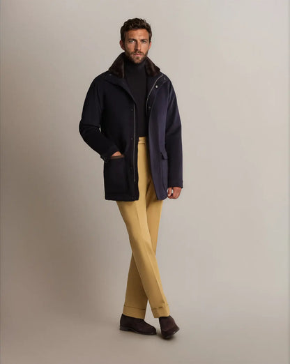 A man wearing a Cavour Navy Winter Car Coat with Removable Cashmere Collar over a black turtleneck stands against a grey background, paired with light yellow trousers and dark brown suede shoes. , available at Richard Gelding Mayfair