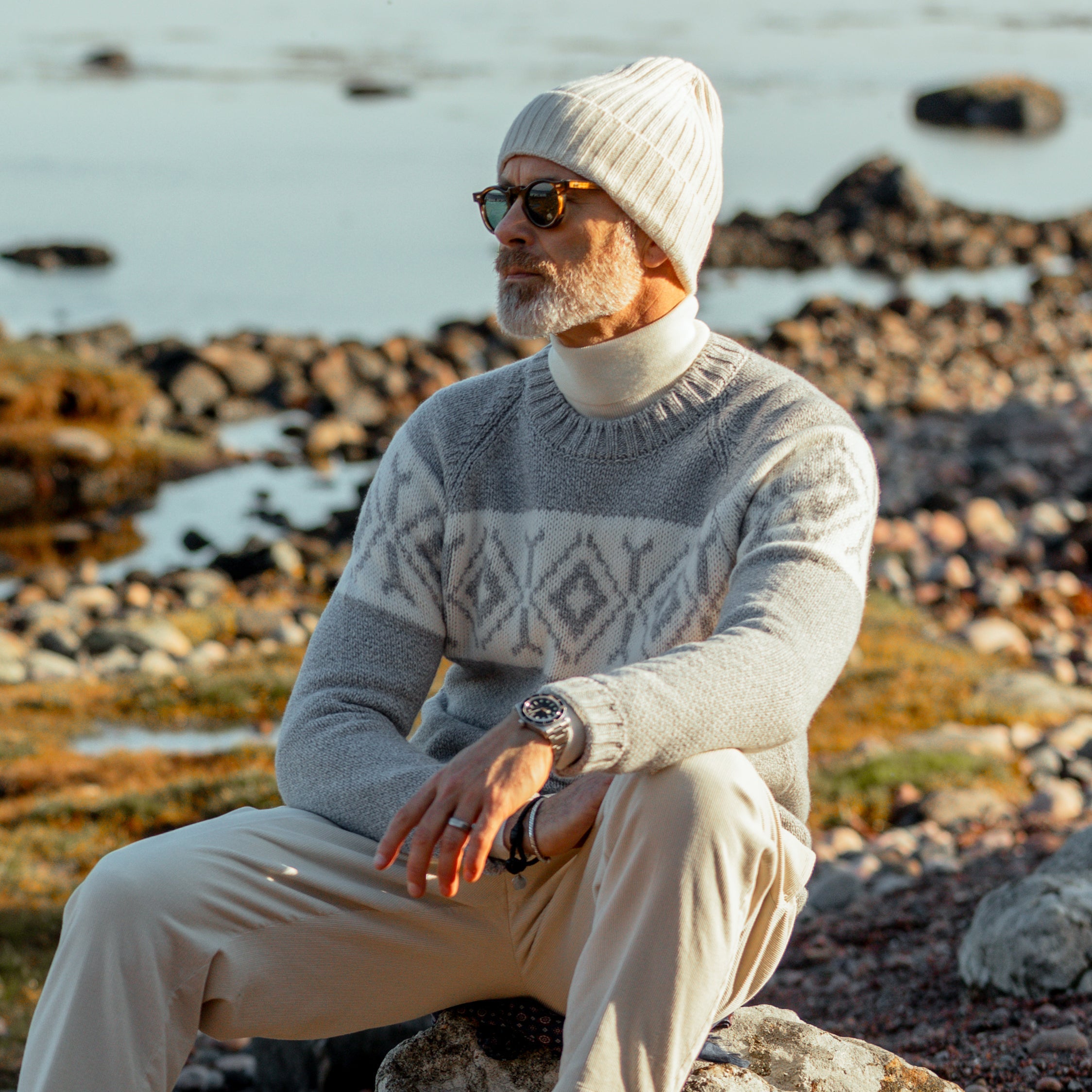A man wearing sunglasses, a Grey Eco Cashmere Norwegian Crewneck Sweater by Gran Sasso, and a beanie sits on rocks by a body of water, embodying a touch of Norwegian design with his calm expression.