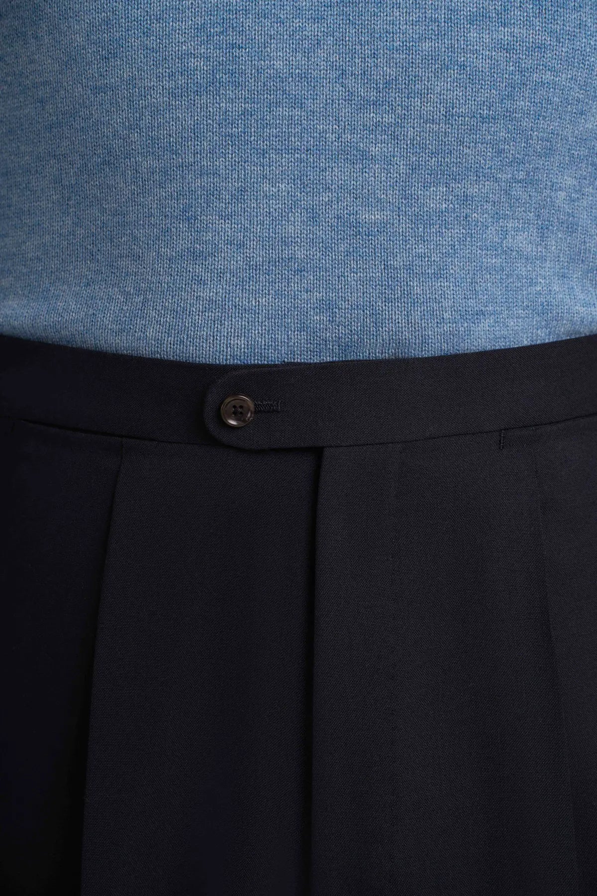 Cavour Navy Blue Wide Leg Pleated Twist Wool Trousers in a tailored fit are worn with a light blue knit sweater, featuring a high-waisted silhouette, a single dark button closure, and deep vertical pleats falling from the waist. , available at Richard Gelding Mayfair
