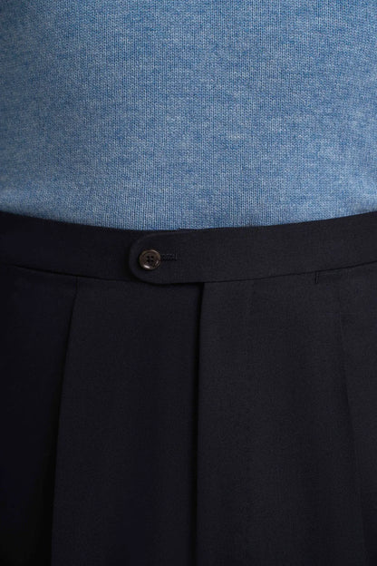 Cavour Navy Blue Wide Leg Pleated Twist Wool Trousers in a tailored fit are worn with a light blue knit sweater, featuring a high-waisted silhouette, a single dark button closure, and deep vertical pleats falling from the waist. , available at Richard Gelding Mayfair