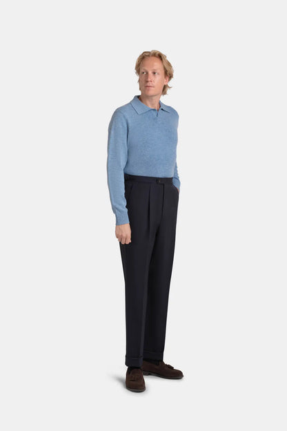 A man with wavy blond hair stands against a plain white background wearing a light blue long-sleeved polo shirt tucked into Cavour Navy Blue Wide Leg Pleated Twist Wool Trousers with brown suede loafers. , available at Richard Gelding Mayfair