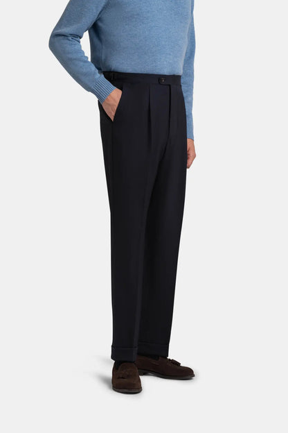 A person wears Navy Blue Wide Leg Pleated Twist Wool Trousers by Cavour in a wide-leg tailored fit with a pleated waist and cuffed hems, paired with a light blue long-sleeved sweater and dark brown suede tassel loafers against a plain light gray background. , available at Richard Gelding Mayfair