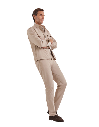 A man stands with arms crossed, smiling slightly and facing left, wearing Canali Beige Cotton Poplin Self-Belt Trouser and a matching beige jacket, paired with dark brown shoes, set against a plain white background. , available at Richard Gelding Mayfair