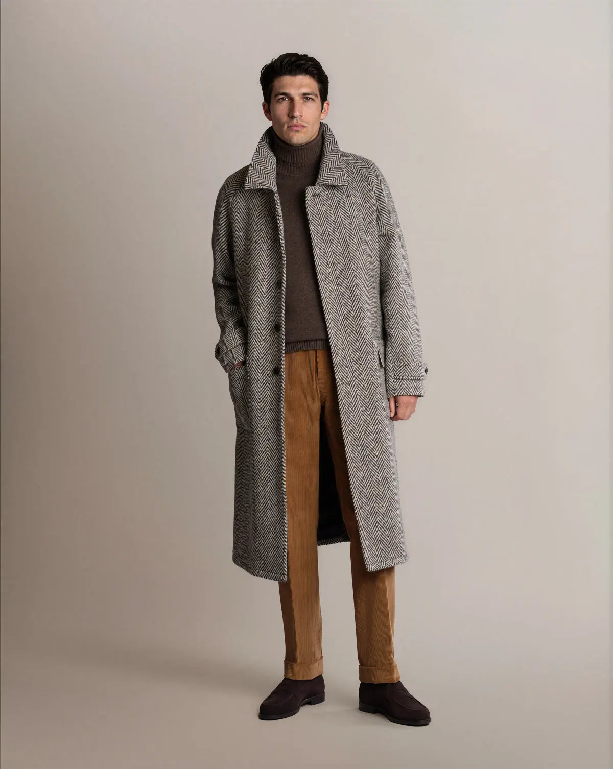 A man stands facing forward wearing a Cavour Black Raglan Tweed Coat in grey herringbone over a brown turtleneck sweater and tan corduroy trousers with dark brown suede shoes against a light grey studio background. , available at Richard Gelding Mayfair