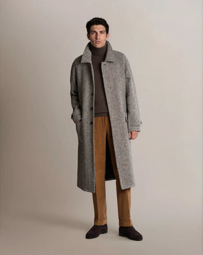 A man stands facing forward wearing a Cavour Black Raglan Tweed Coat in grey herringbone over a brown turtleneck sweater and tan corduroy trousers with dark brown suede shoes against a light grey studio background. , available at Richard Gelding Mayfair