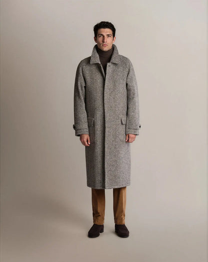 A man stands centered against a neutral grey background wearing a grey herringbone Cavour Black Raglan Tweed Coat over a brown turtleneck, tan trousers, and dark brown suede shoes; the coat features a tall collar, side pockets with flaps, and buttoned cuff straps. , available at Richard Gelding Mayfair