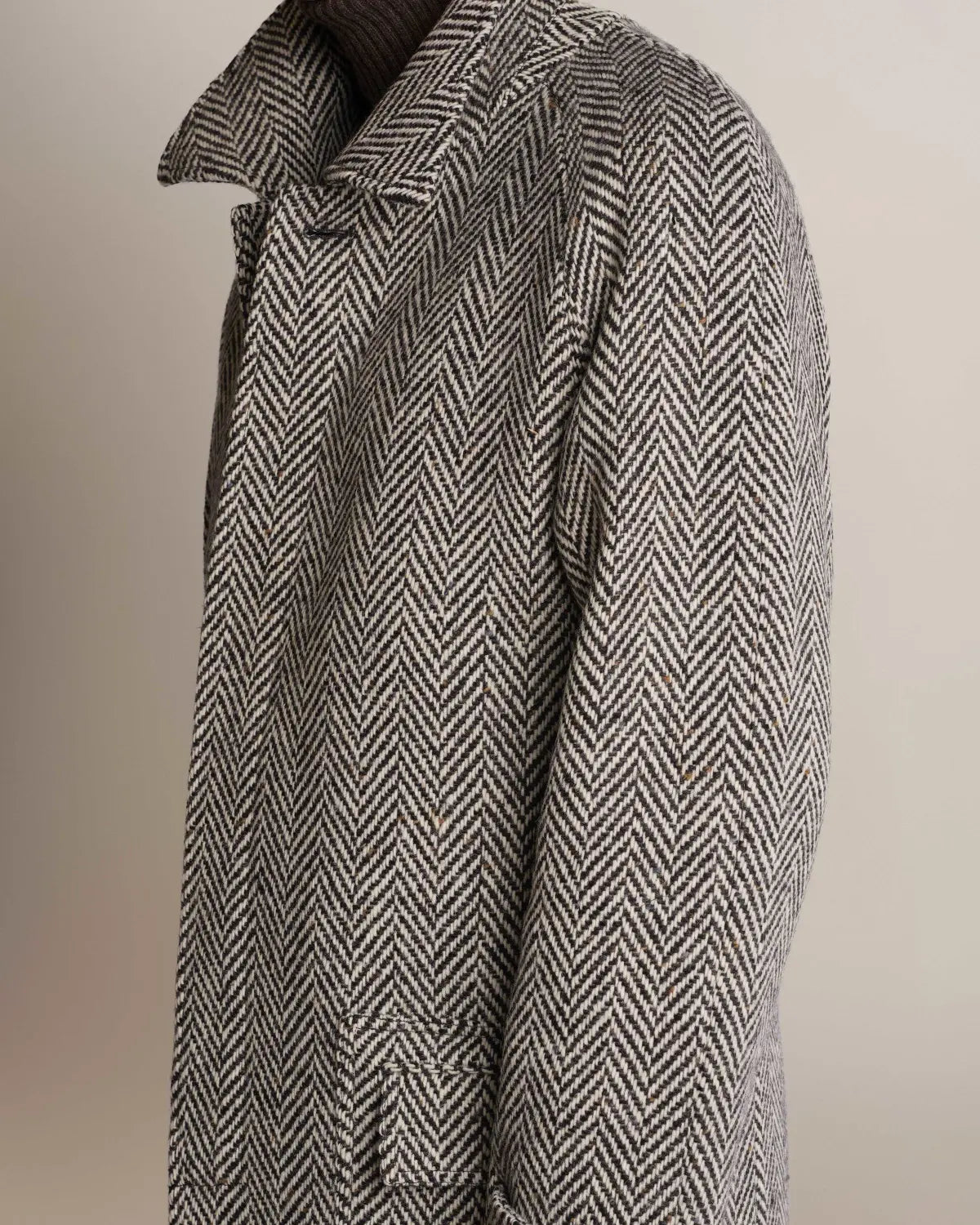 A side profile of a person wearing a Cavour Black Raglan Tweed Coat in a black and white herringbone pattern with raglan sleeves, a wide collar, and a dark knit turtleneck underneath against a plain light background. , available at Richard Gelding Mayfair
