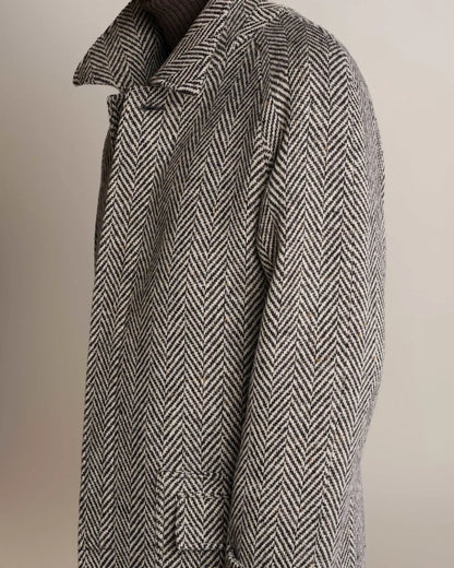 A side profile of a person wearing a Cavour Black Raglan Tweed Coat in a black and white herringbone pattern with raglan sleeves, a wide collar, and a dark knit turtleneck underneath against a plain light background. , available at Richard Gelding Mayfair