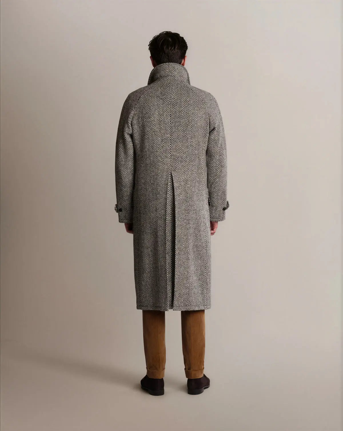 A man seen from behind wears a Cavour Black Raglan Tweed Coat, a knee-length, grey and black herringbone piece with raglan sleeves, a high collar, and a deep single back vent, paired with tan corduroy trousers and dark brown shoes against a plain light background. , available at Richard Gelding Mayfair