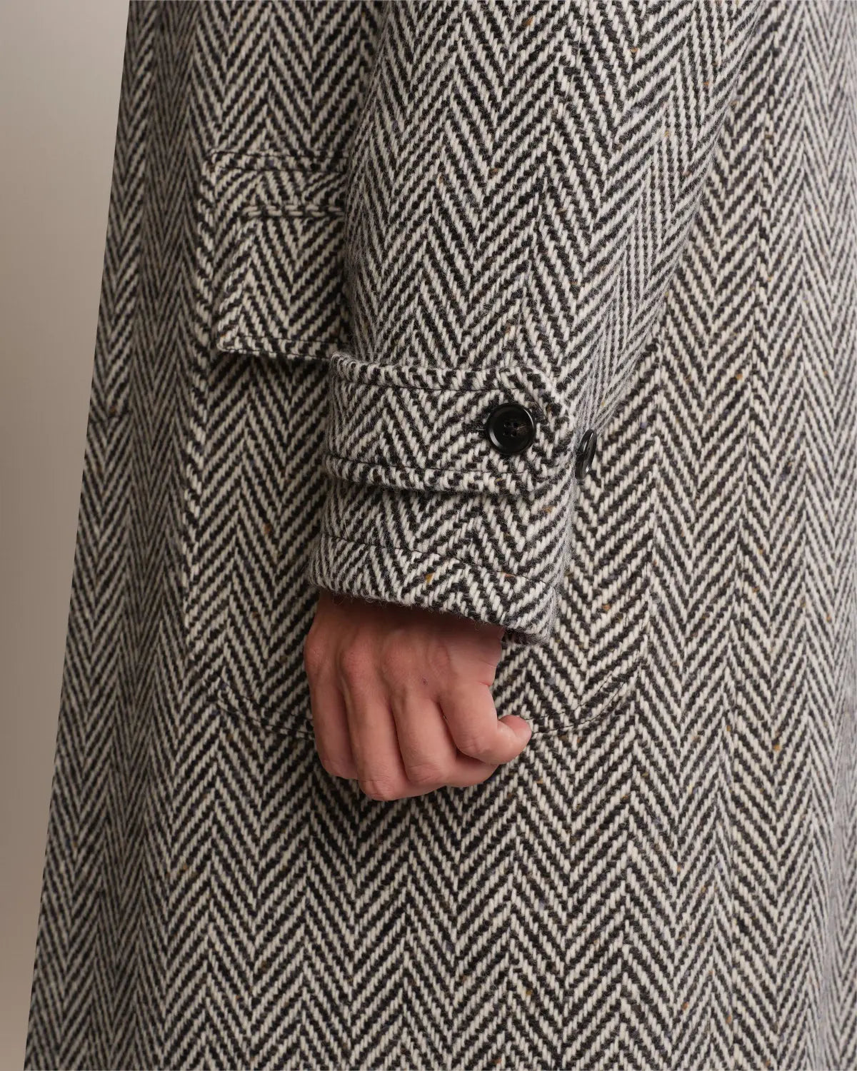 A close-up of a Cavour Black Raglan Tweed Coat featuring a black and white herringbone pattern, raglan sleeves with buttoned cuff straps, and a waist flap pocket, with a hand partially visible at the side. , available at Richard Gelding Mayfair