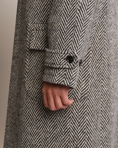 A close-up of a Cavour Black Raglan Tweed Coat featuring a black and white herringbone pattern, raglan sleeves with buttoned cuff straps, and a waist flap pocket, with a hand partially visible at the side. , available at Richard Gelding Mayfair