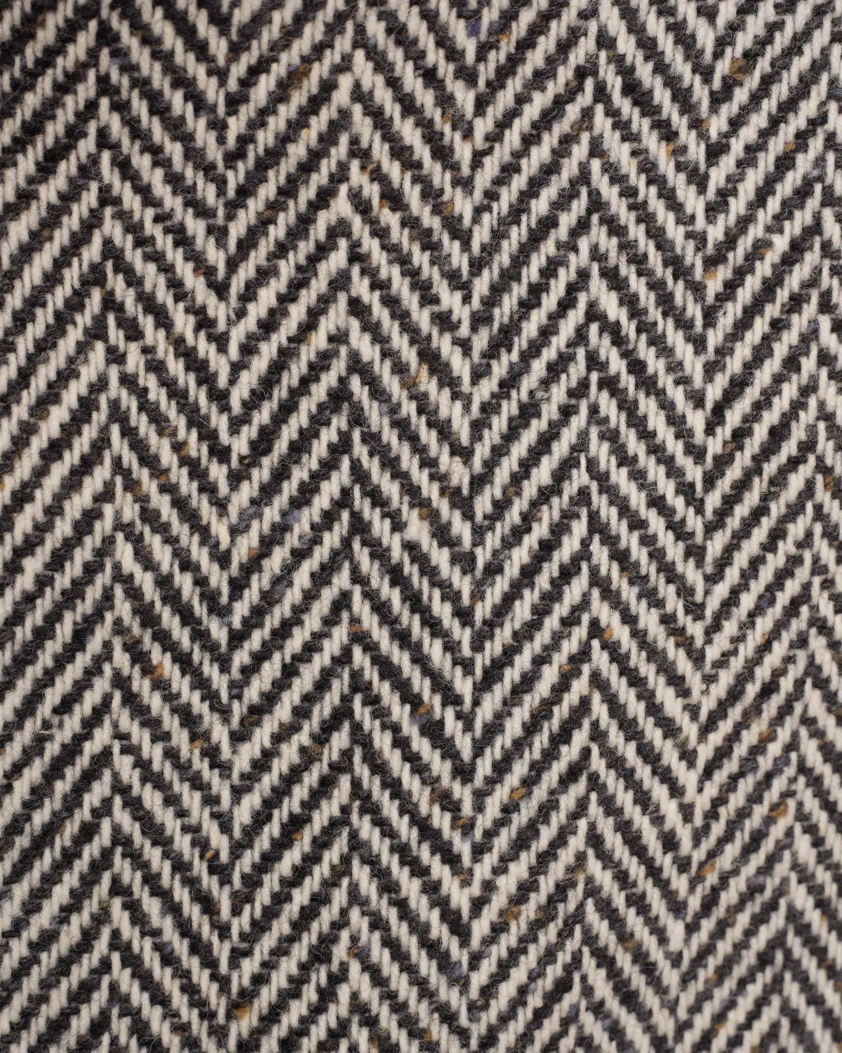 A close-up of the Black Raglan Tweed Coat in heavy wool with a black and white herringbone pattern, featuring subtle flecks of brown and blue woven throughout the textured fabric. , available at Richard Gelding Mayfair