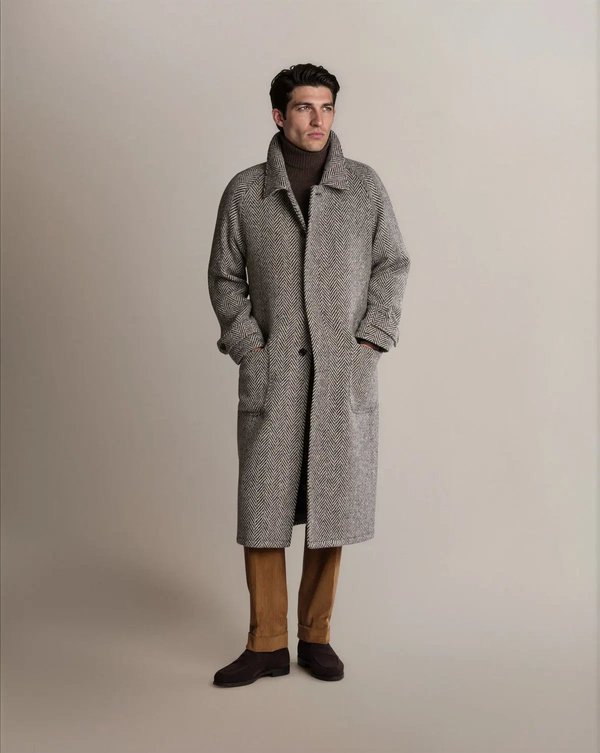 A man stands wearing a Cavour Black Raglan Tweed Coat in grey herringbone with raglan sleeves over a dark brown turtleneck, tan corduroy trousers, and dark brown suede loafers, looking off to the side against a light grey background. , available at Richard Gelding Mayfair
