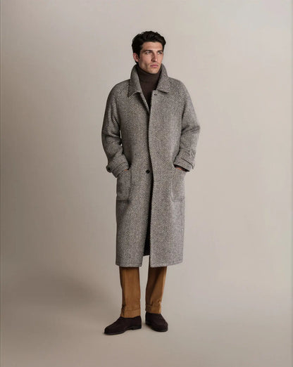 A man stands wearing a Cavour Black Raglan Tweed Coat in grey herringbone with raglan sleeves over a dark brown turtleneck, tan corduroy trousers, and dark brown suede loafers, looking off to the side against a light grey background. , available at Richard Gelding Mayfair