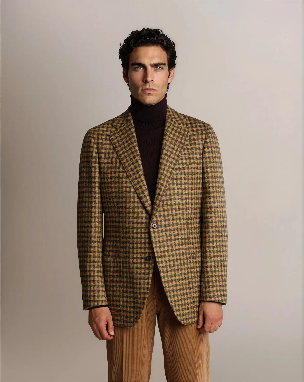 A man with dark hair wears a brown and green Cavour Brown Shepherds Check Tweed Blazer over a dark brown turtleneck, paired with tan corduroy trousers against a plain, light grey background. , available at Richard Gelding Mayfair