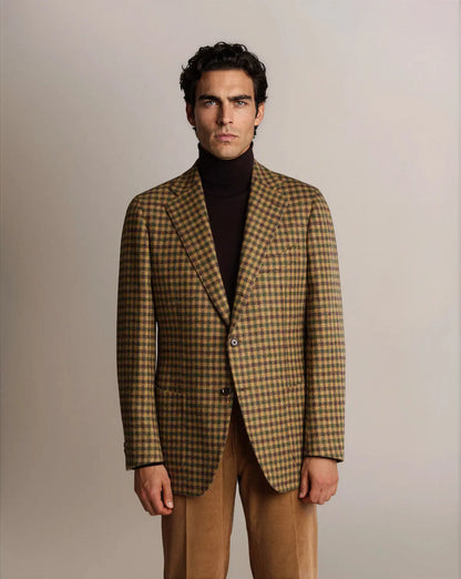 A man with dark hair wears a brown and green Cavour Brown Shepherds Check Tweed Blazer over a dark brown turtleneck, paired with tan corduroy trousers against a plain, light grey background. , available at Richard Gelding Mayfair