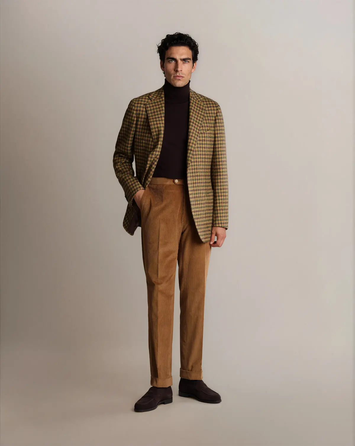 A man with dark hair wears a Brown Shepherds Check Tweed Blazer by Cavour over a dark brown turtleneck, paired with light brown corduroy trousers and dark brown suede shoes, standing against a neutral background with one hand in his pocket. , available at Richard Gelding Mayfair