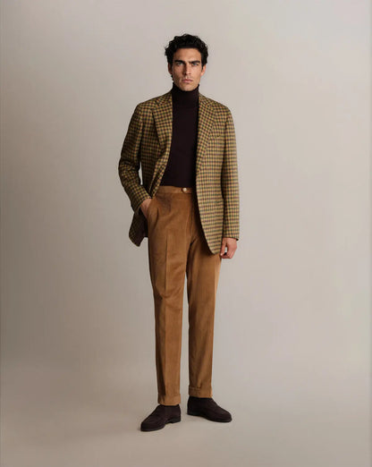 A man with dark hair wears a Brown Shepherds Check Tweed Blazer by Cavour over a dark brown turtleneck, paired with light brown corduroy trousers and dark brown suede shoes, standing against a neutral background with one hand in his pocket. , available at Richard Gelding Mayfair