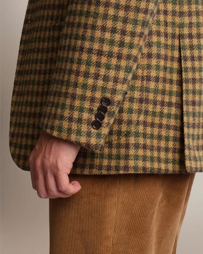 A close-up of a person wearing a Brown Shepherds Check Tweed Blazer by Cavour in brown, green, and tan, featuring four dark buttons at the cuff and tan corduroy trousers against a plain light background. , available at Richard Gelding Mayfair